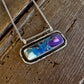 A silver-toned chain necklace with a rectangular bezel pendant featuring a marbled inlay of blue, purple, white, and gold, displayed on a rustic dark wood surface.