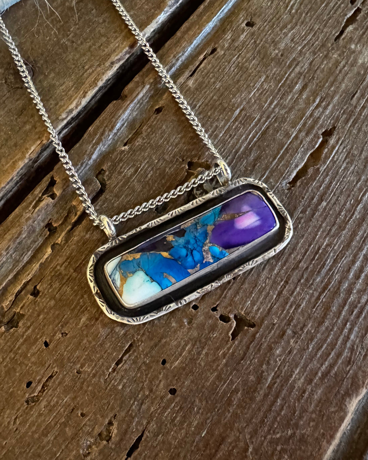 A silver-toned chain necklace with a rectangular bezel pendant featuring a marbled inlay of blue, purple, white, and gold, displayed on a rustic dark wood surface.