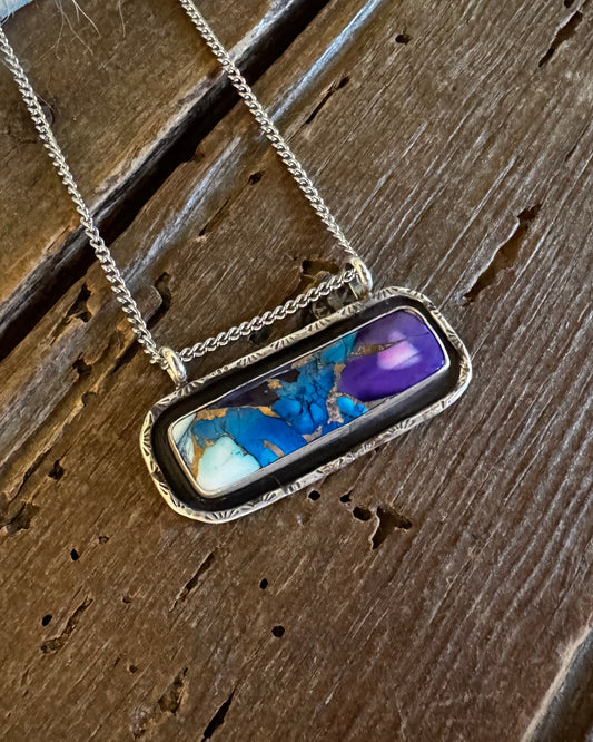 A silver-toned chain necklace with a rectangular bezel pendant featuring a marbled inlay of blue, purple, white, and gold, displayed on a rustic dark wood surface.