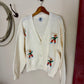 White cardigan with embroidered designs on a hanger against a plain wall.