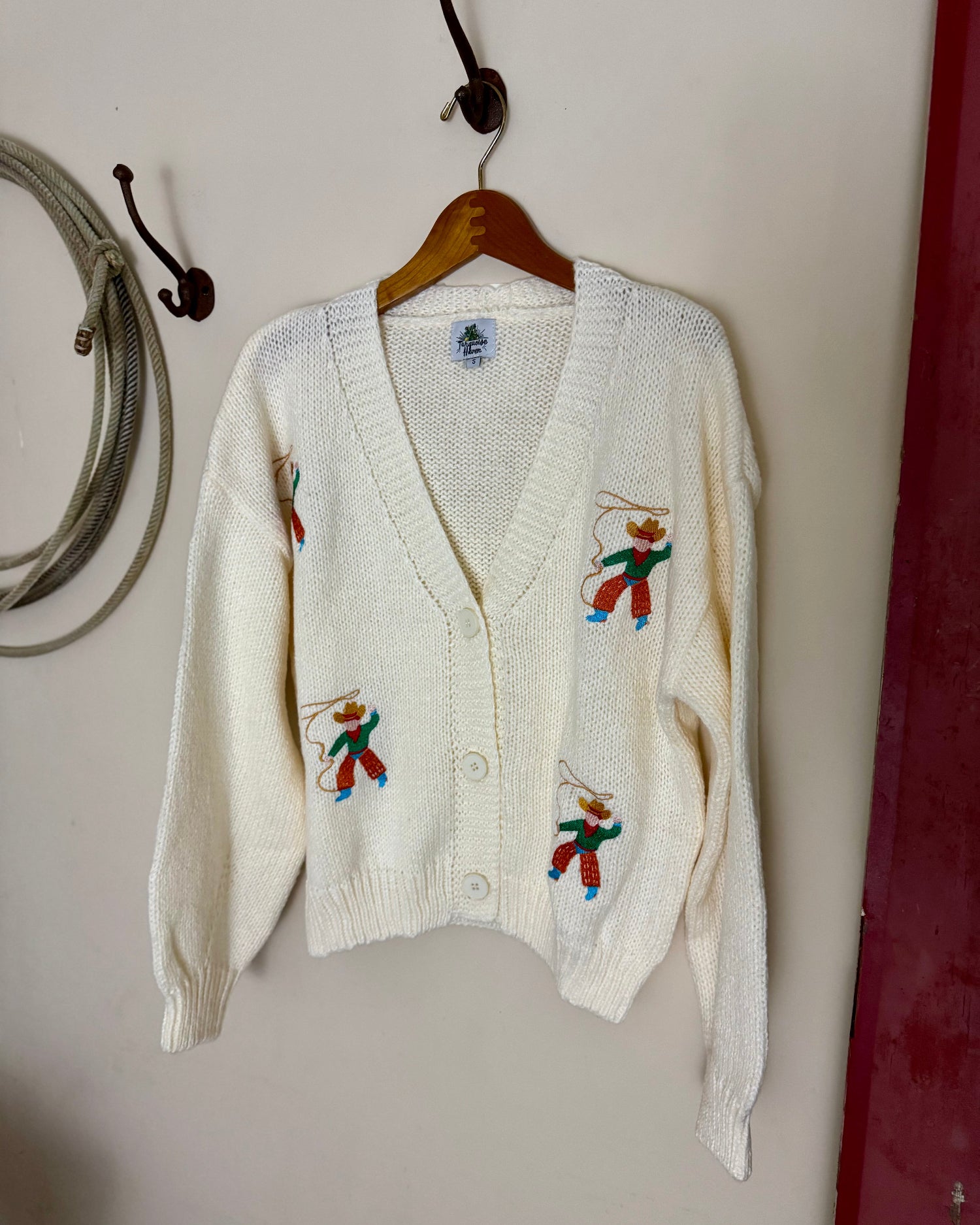White cardigan with embroidered designs on a hanger against a plain wall.
