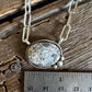 A silver-toned necklace with a textured, organic-shaped stone pendant is displayed on a rustic wooden surface next to a metal ruler for scale.