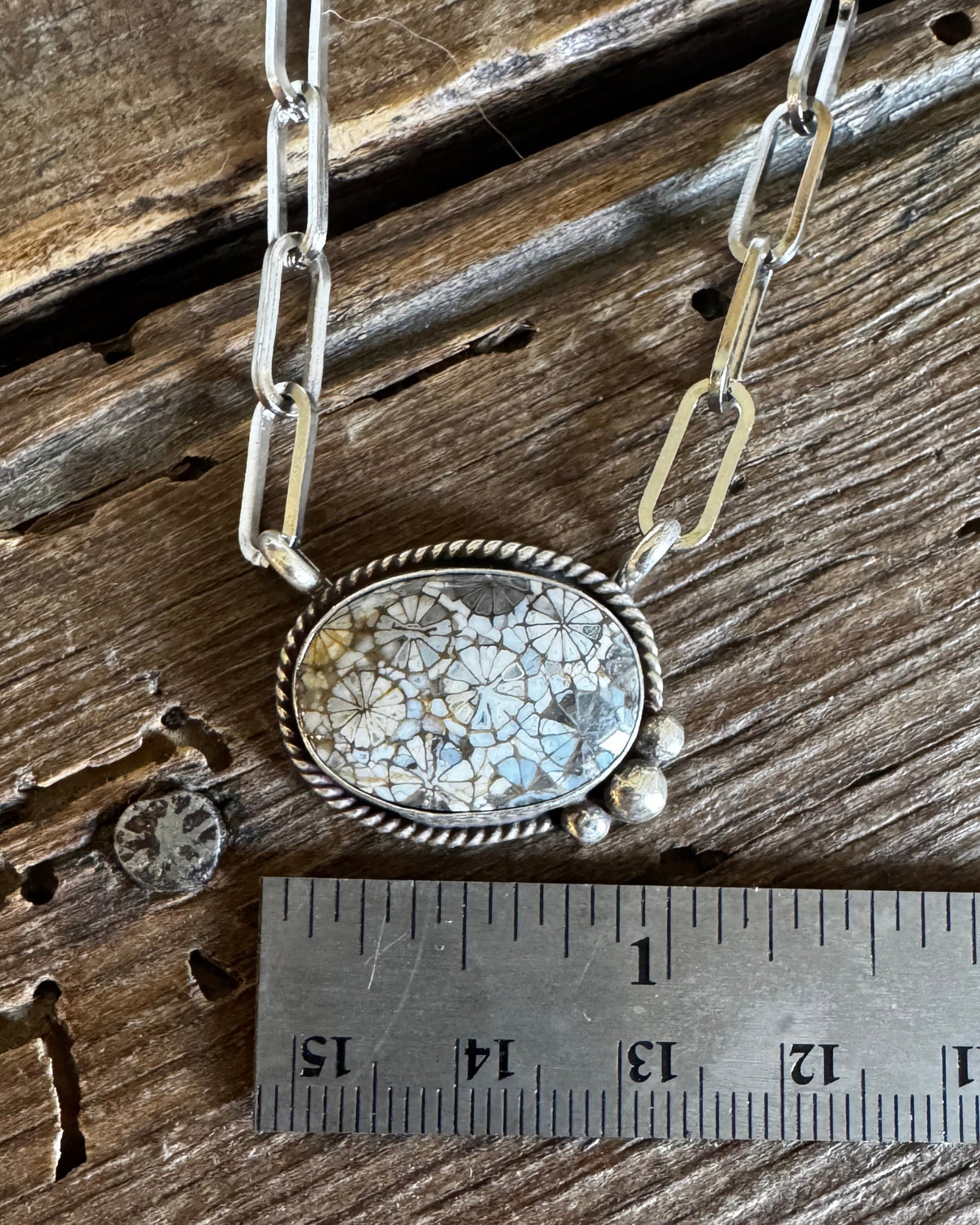 A silver-toned necklace with a textured, organic-shaped stone pendant is displayed on a rustic wooden surface next to a metal ruler for scale.