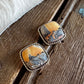 A pair of square stud earrings with twisted silver-tone borders and inlay stones showing a landscape scene in orange, brown, and gray, resting on a rustic wooden surface next to white fur.