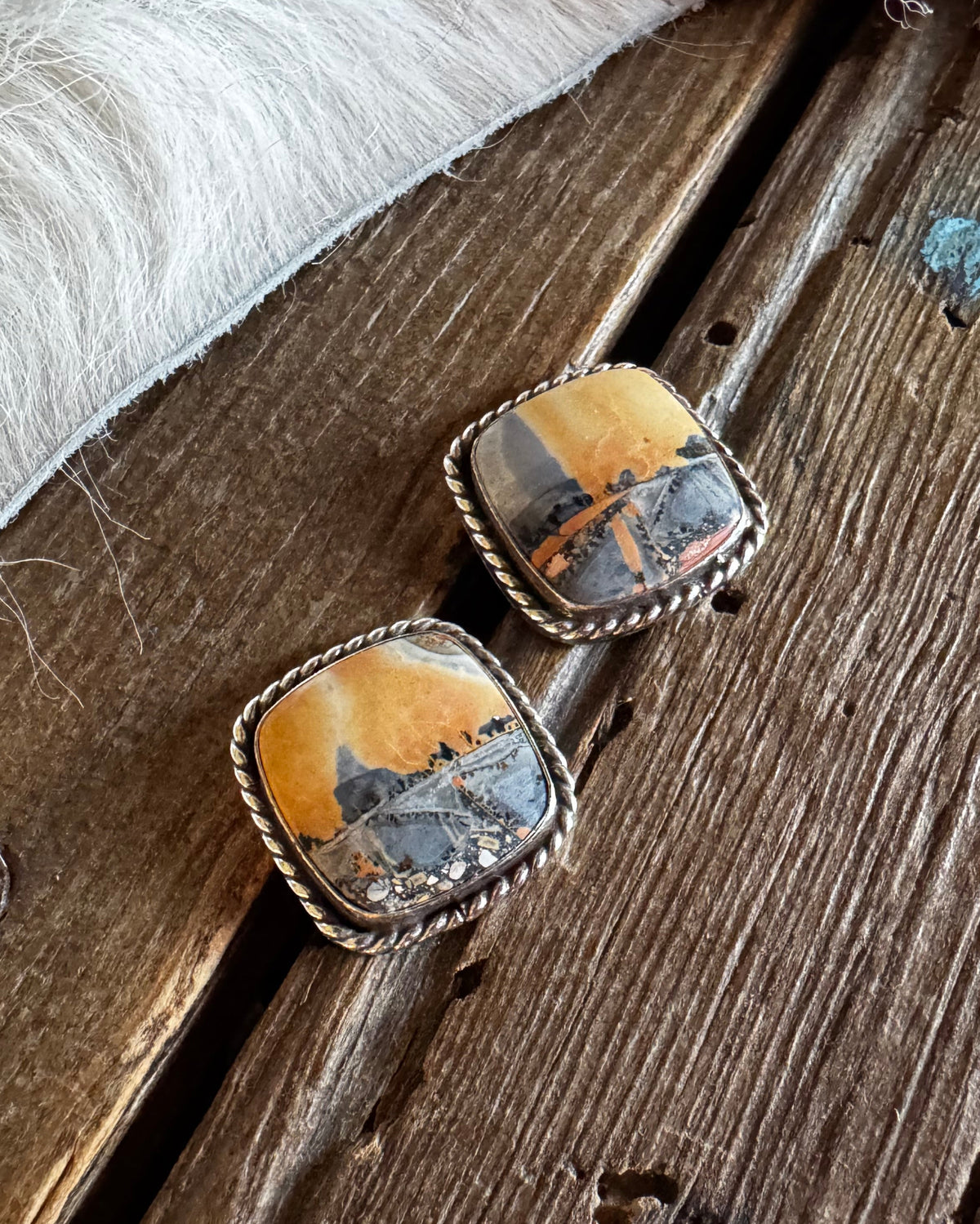 A pair of square stud earrings with twisted silver-tone borders and inlay stones showing a landscape scene in orange, brown, and gray, resting on a rustic wooden surface next to white fur.
