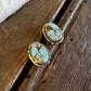 Two oval turquoise and gold stud earrings resting on a rustic wooden surface next to a white faux fur trim.