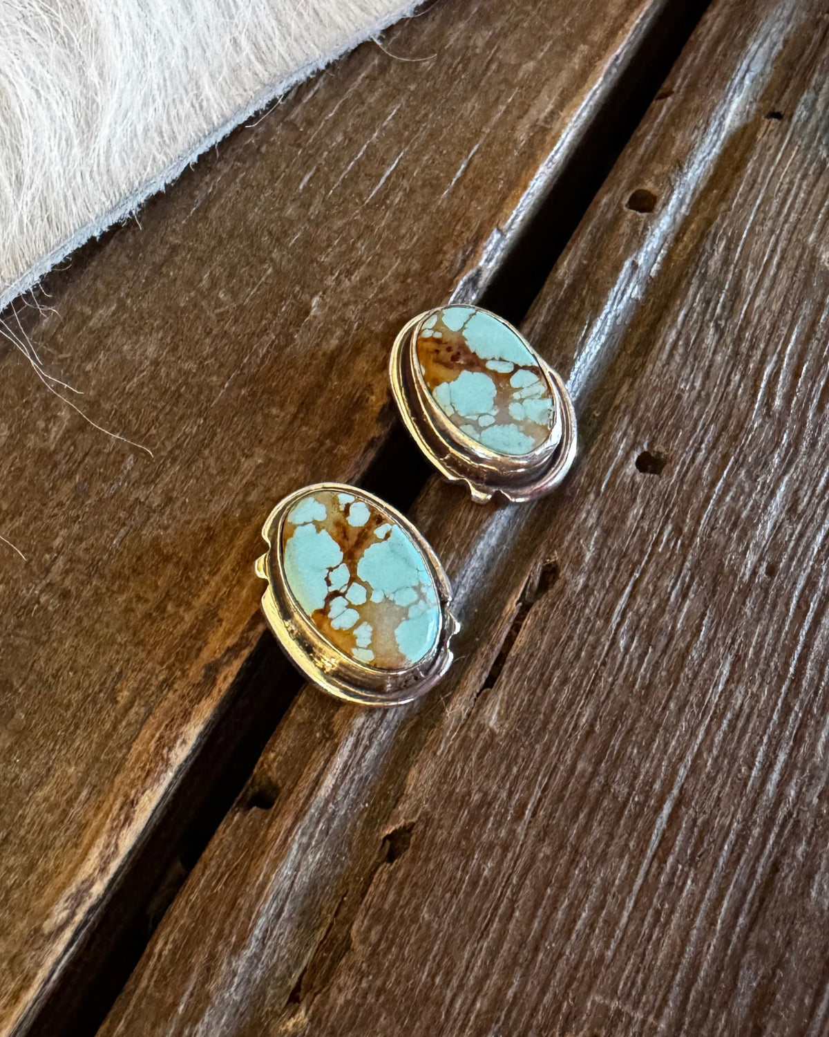 Two oval turquoise and gold stud earrings resting on a rustic wooden surface next to a white faux fur trim.