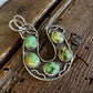 A silver horse shoe pendant featuring five green and yellow marbled cabochon gemstones, displayed on a rustic dark wood background.