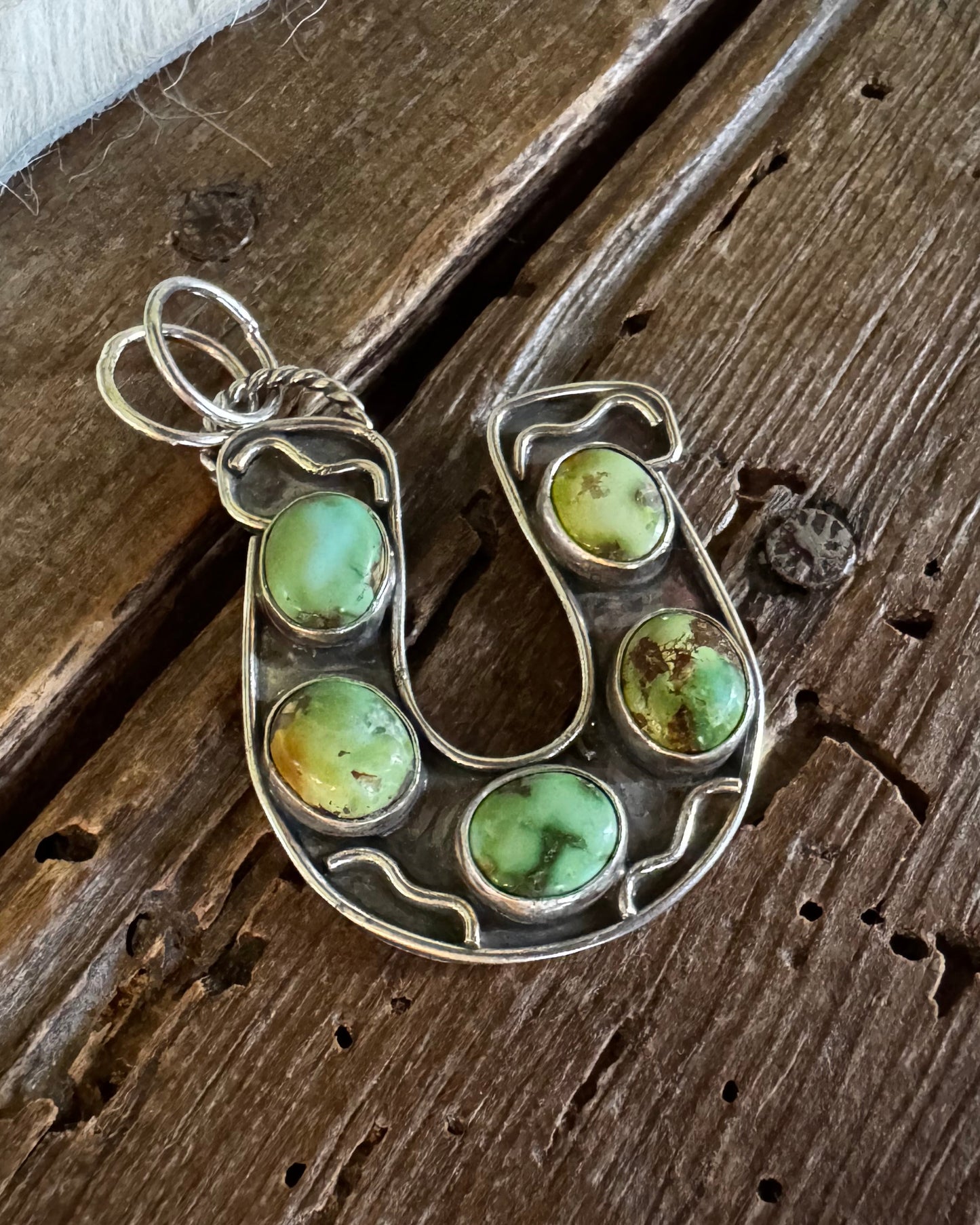 A silver horse shoe pendant featuring five green and yellow marbled cabochon gemstones, displayed on a rustic dark wood background.