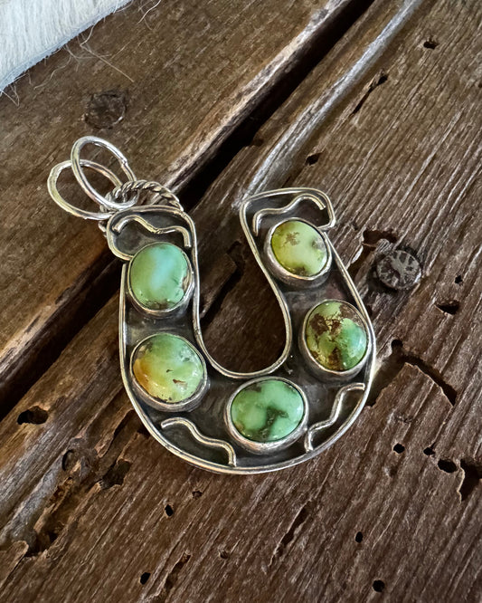 A silver horse shoe pendant featuring five green and yellow marbled cabochon gemstones, displayed on a rustic dark wood background.