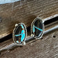 A pair of oval silver stud earrings with turquoise inlay, resting on a rustic wooden surface.