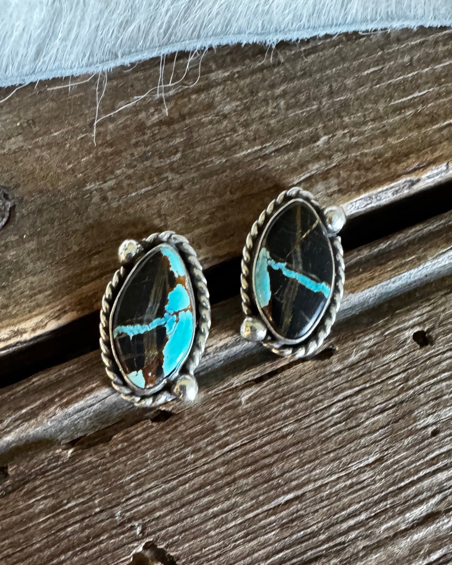 A pair of oval silver stud earrings with turquoise inlay, resting on a rustic wooden surface.