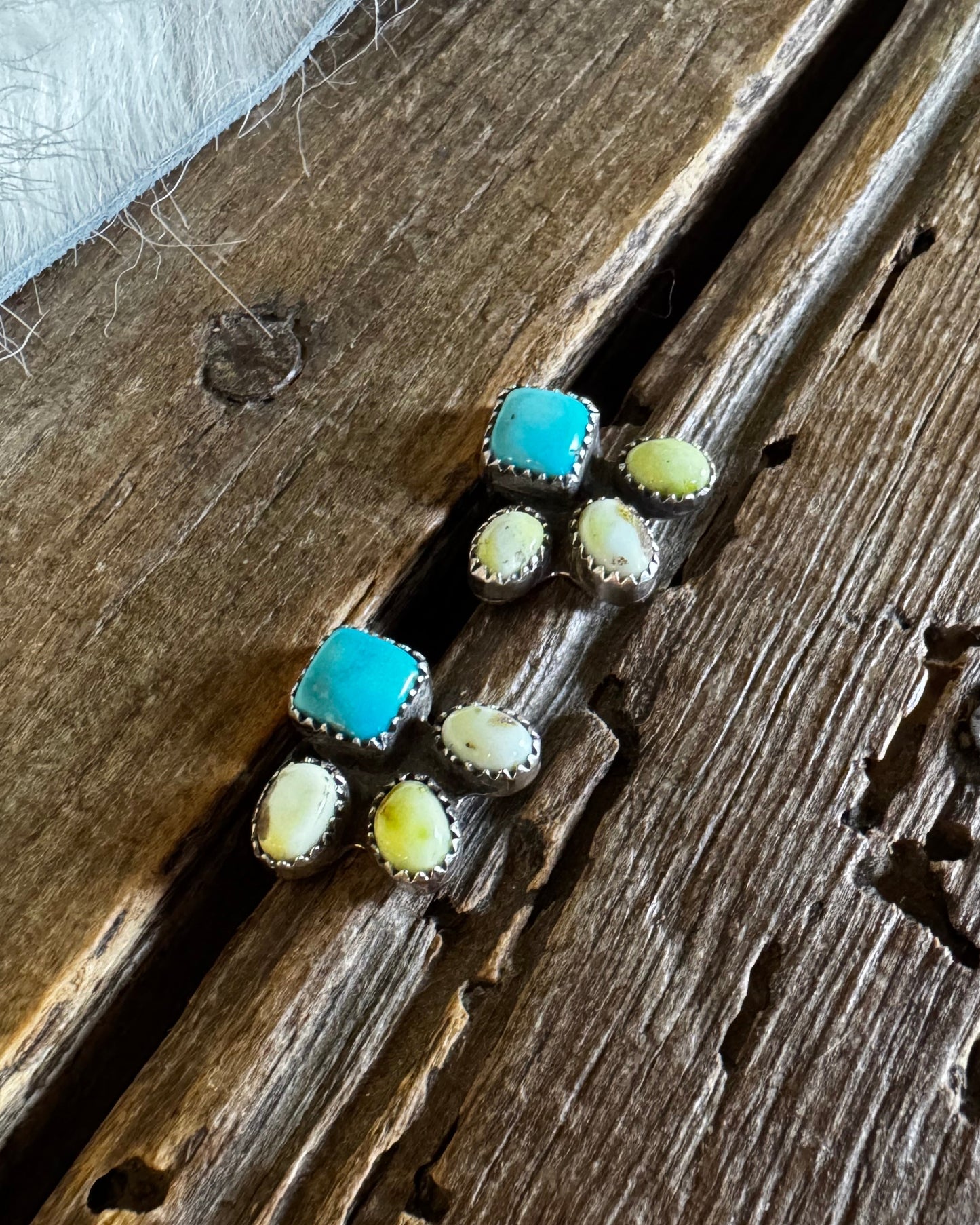 A pair of silver stud earrings with turquoise blue square centers and small cream-colored oval stones, displayed on a rustic wooden surface next to a white faux fur fabric.