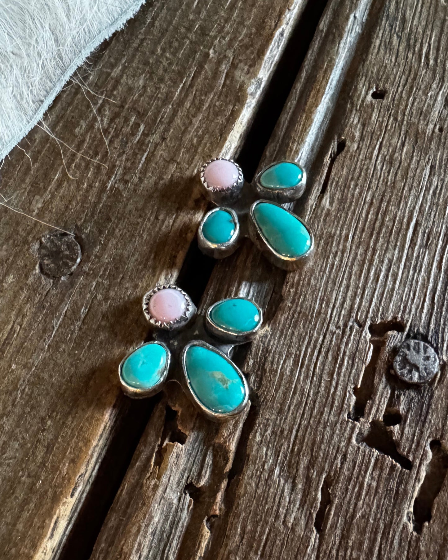 A pair of handmade turquoise and pink stone stud earrings in silver settings, displayed on a rustic wooden surface next to a white fur trim.