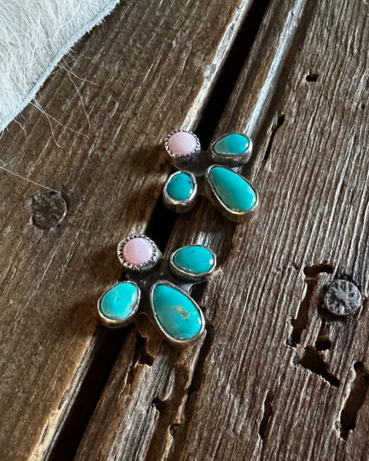 A pair of handmade turquoise and pink stone stud earrings in silver settings, displayed on a rustic wooden surface next to a white fur trim.