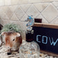 Clear bottle with a horse-shaped stopper on a kitchen counter, next to a cow figurine and a chalkboard with 'COW' written on it.