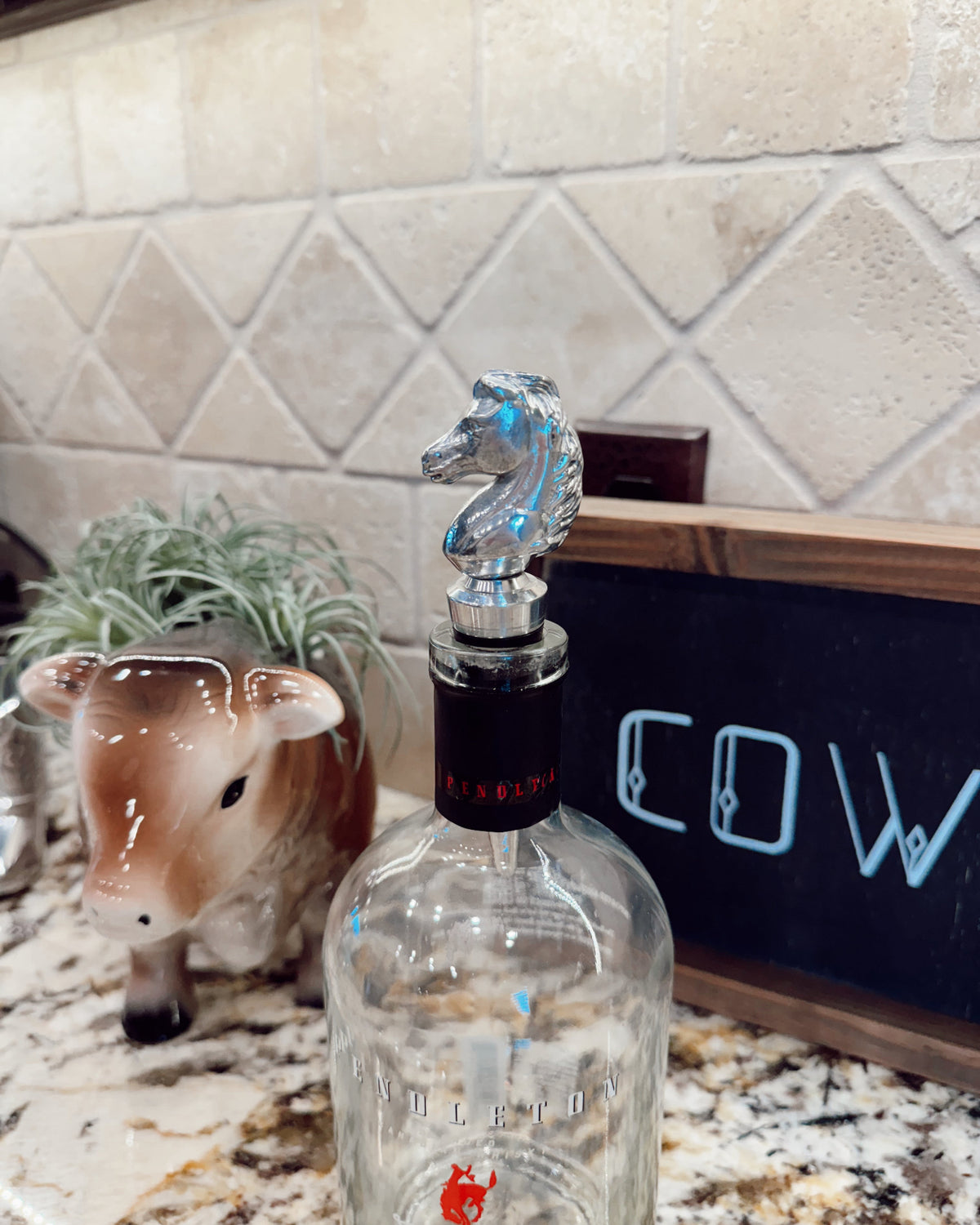 Clear bottle with a horse-shaped stopper on a kitchen counter, next to a cow figurine and a chalkboard with 'COW' written on it.