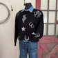 Black sweater with cowboy-themed designs on a mannequin against a rustic background.