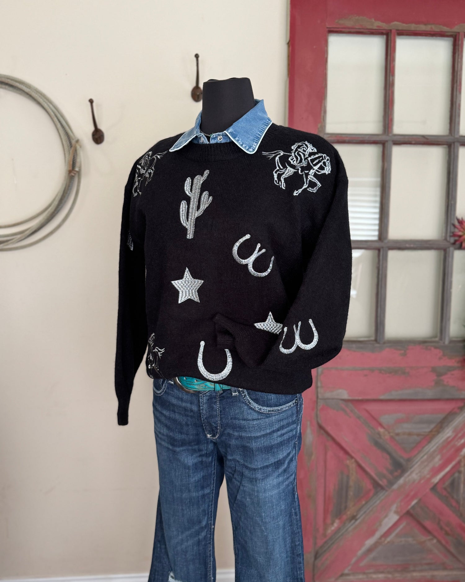 Black sweater with cowboy-themed designs on a mannequin against a rustic background.