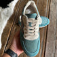 A teal and beige low-top sneaker held up by a hand over a rustic wooden floor, with a second matching sneaker blurred in the background.