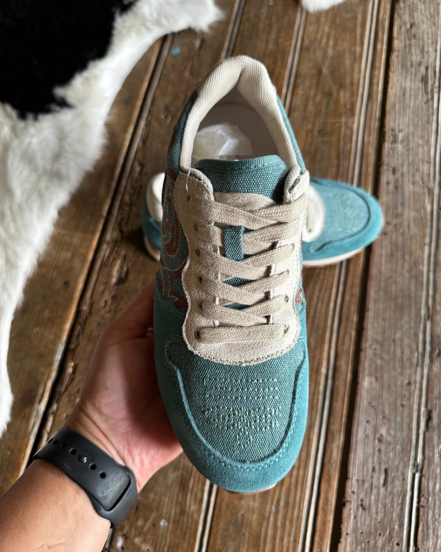 A teal and beige low-top sneaker held up by a hand over a rustic wooden floor, with a second matching sneaker blurred in the background.