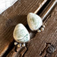 A pair of handmade sterling silver earrings featuring light green and white marbled stone pendants with small silver bead drops, resting on a rustic dark wood surface next to a white fur trim.