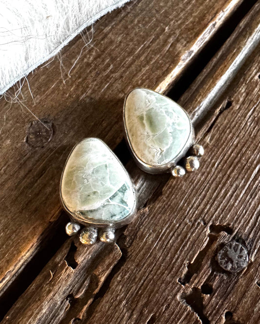 A pair of handmade sterling silver earrings featuring light green and white marbled stone pendants with small silver bead drops, resting on a rustic dark wood surface next to a white fur trim.