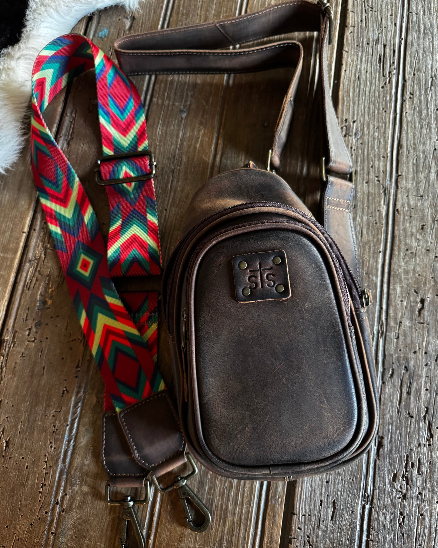 A dark brown leather crossbody bag with a colorful geometric patterned strap, displayed on a rustic wooden surface.