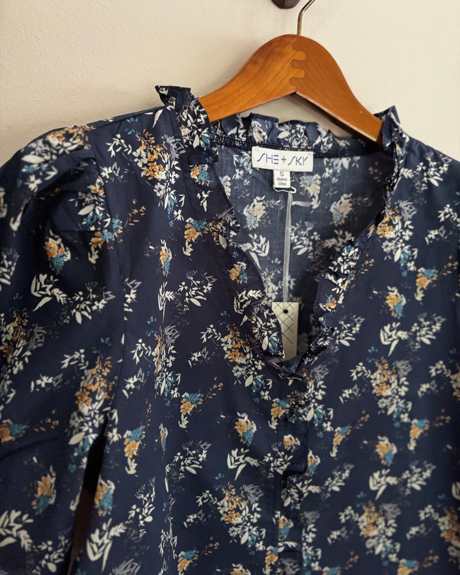 Navy floral blouse on a hanger with a visible brand tag against a beige wall.