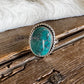 A silver ring featuring a round turquoise stone, displayed on a rustic wooden surface next to a white faux fur trim.