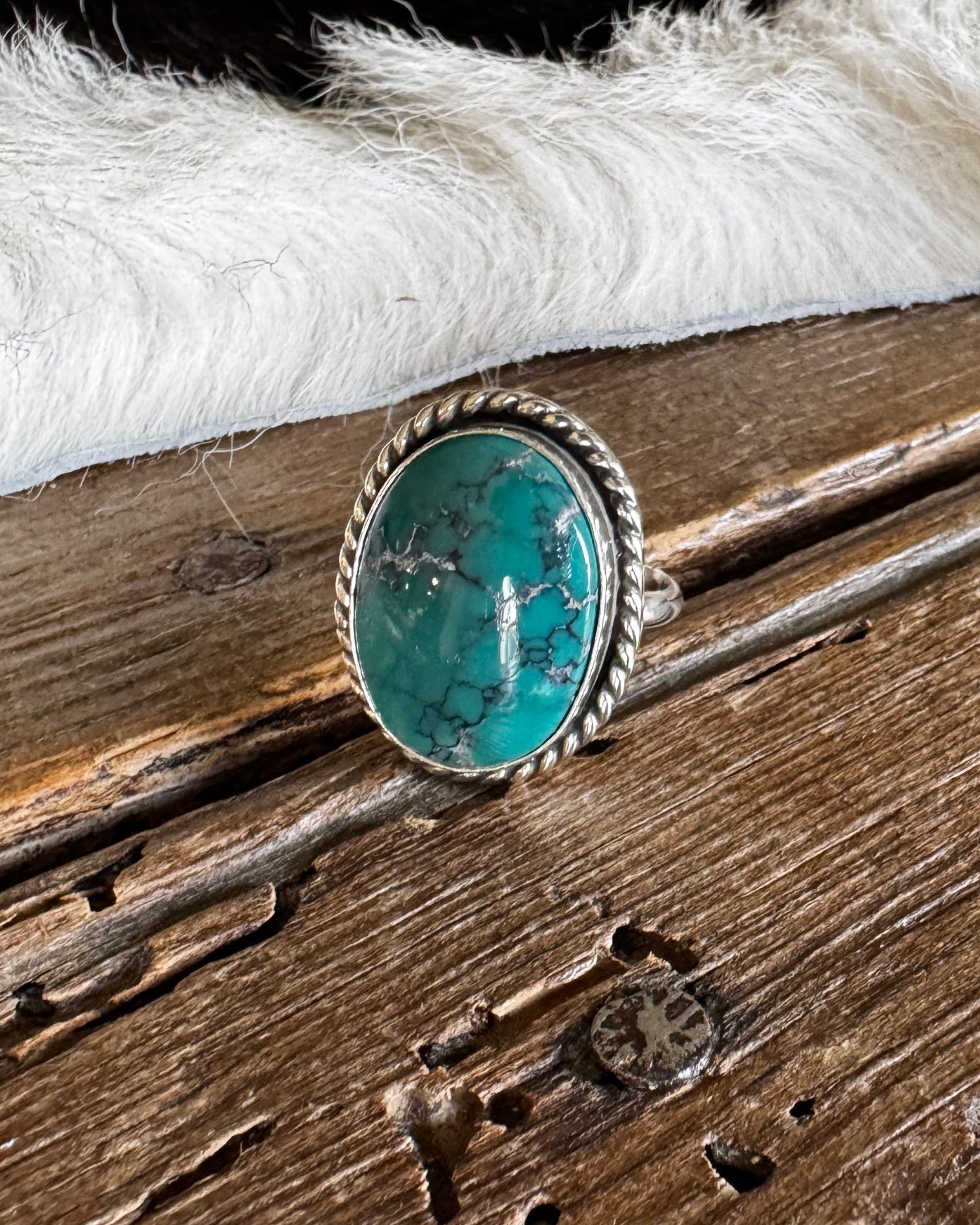 A silver ring featuring a round turquoise stone, displayed on a rustic wooden surface next to a white faux fur trim.