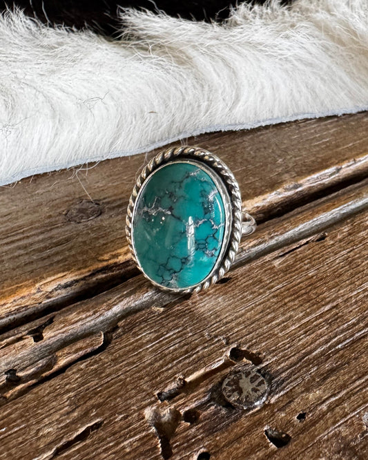A silver ring featuring a round turquoise stone, displayed on a rustic wooden surface next to a white faux fur trim.