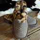 A pair of women's slip-on slippers with a brown felt upper, leopard-print faux fur trim, and gold star-shaped studs, displayed on a rustic wooden surface next to a black and white cowhide rug.