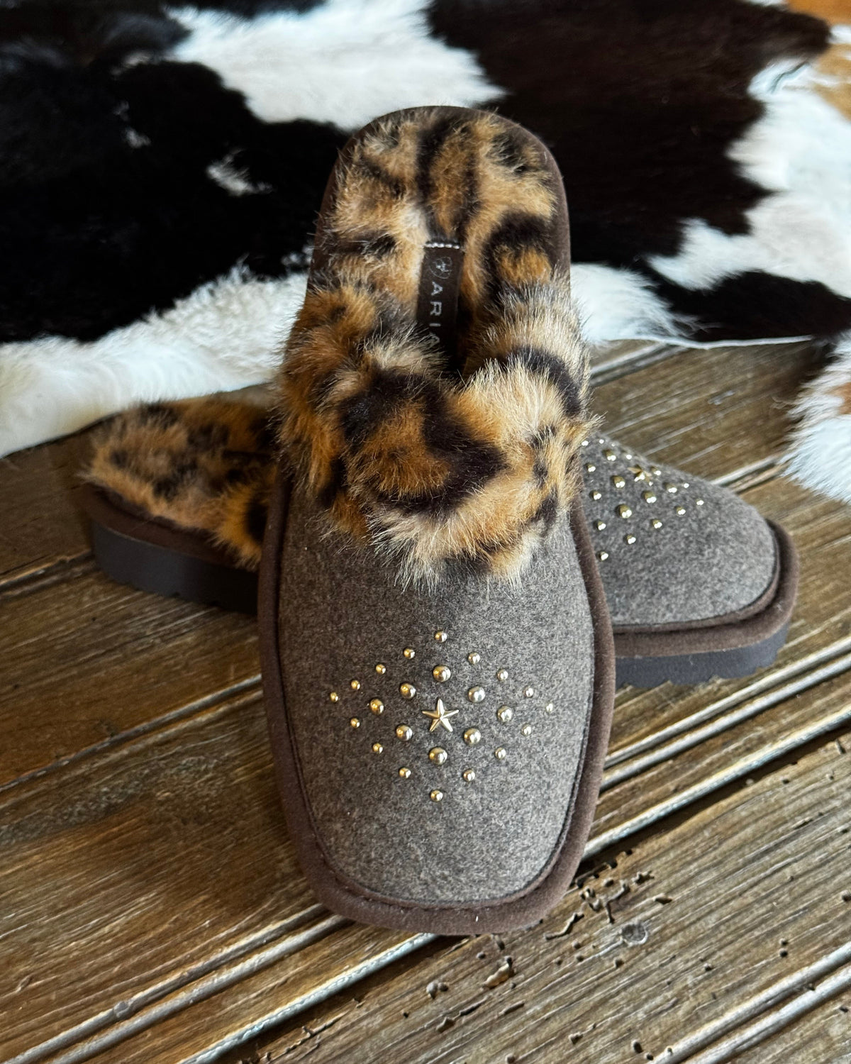 A pair of women's slip-on slippers with a brown felt upper, leopard-print faux fur trim, and gold star-shaped studs, displayed on a rustic wooden surface next to a black and white cowhide rug.