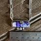 A silver-toned necklace with a rectangular pendant featuring a purple and blue floral pattern on a white base, displayed on a rustic wooden surface next to a metal ruler for scale.