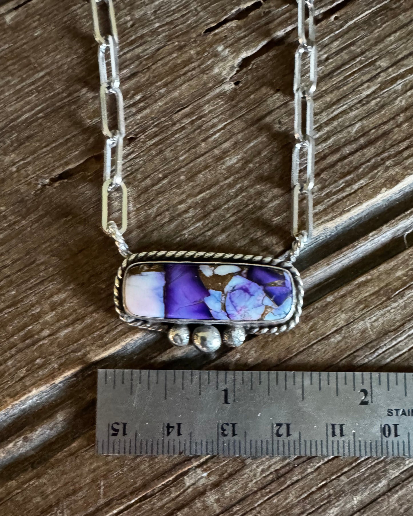 A silver-toned necklace with a rectangular pendant featuring a purple and blue floral pattern on a white base, displayed on a rustic wooden surface next to a metal ruler for scale.