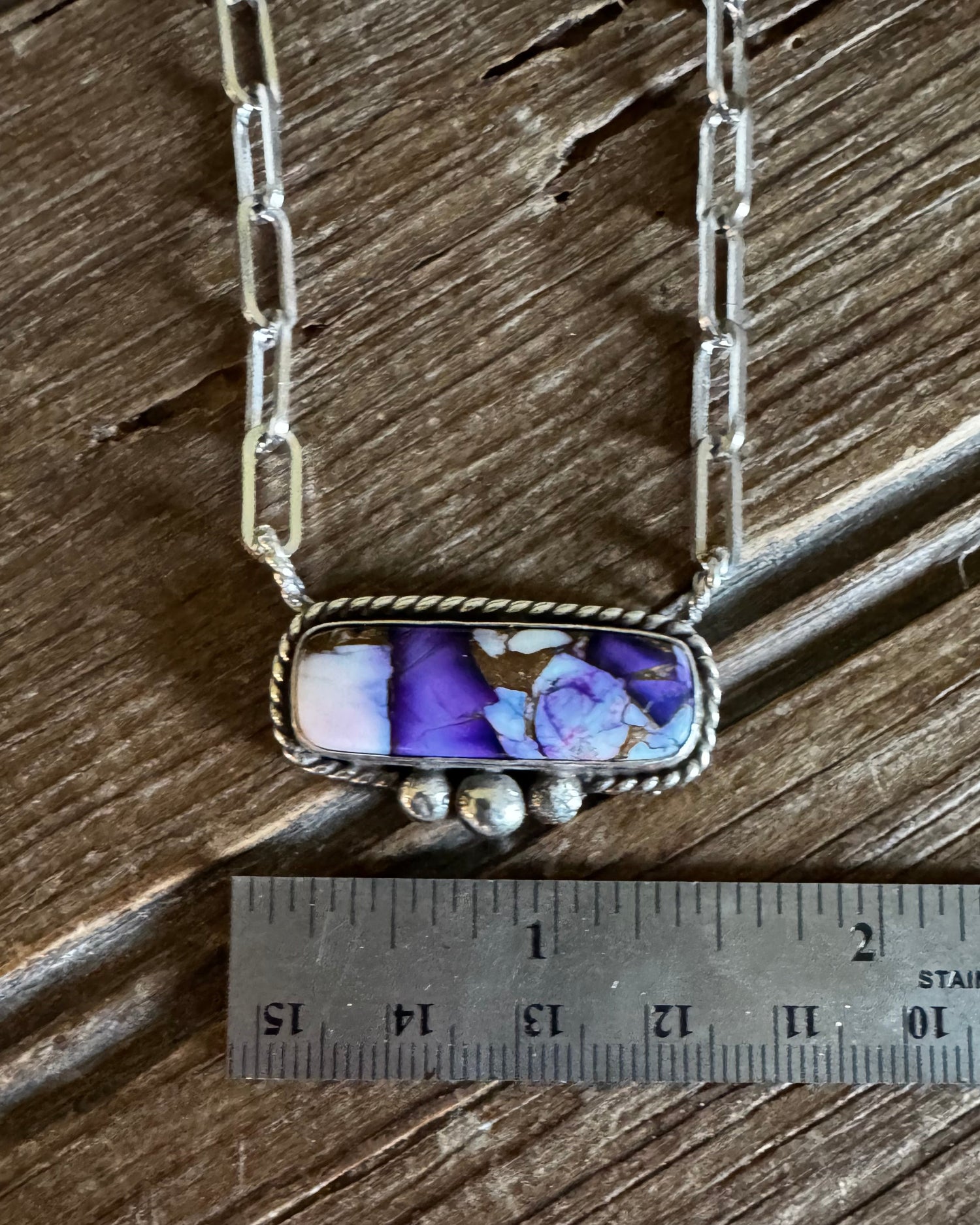 A silver-toned necklace with a rectangular pendant featuring a purple and blue floral pattern on a white base, displayed on a rustic wooden surface next to a metal ruler for scale.