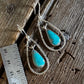 A pair of silver-toned drop earrings with teardrop-shaped turquoise stones, displayed on a rustic wooden surface next to a metal ruler for scale.