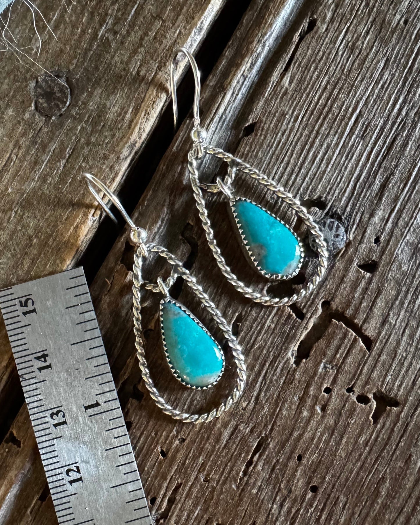 A pair of silver-toned drop earrings with teardrop-shaped turquoise stones, displayed on a rustic wooden surface next to a metal ruler for scale.