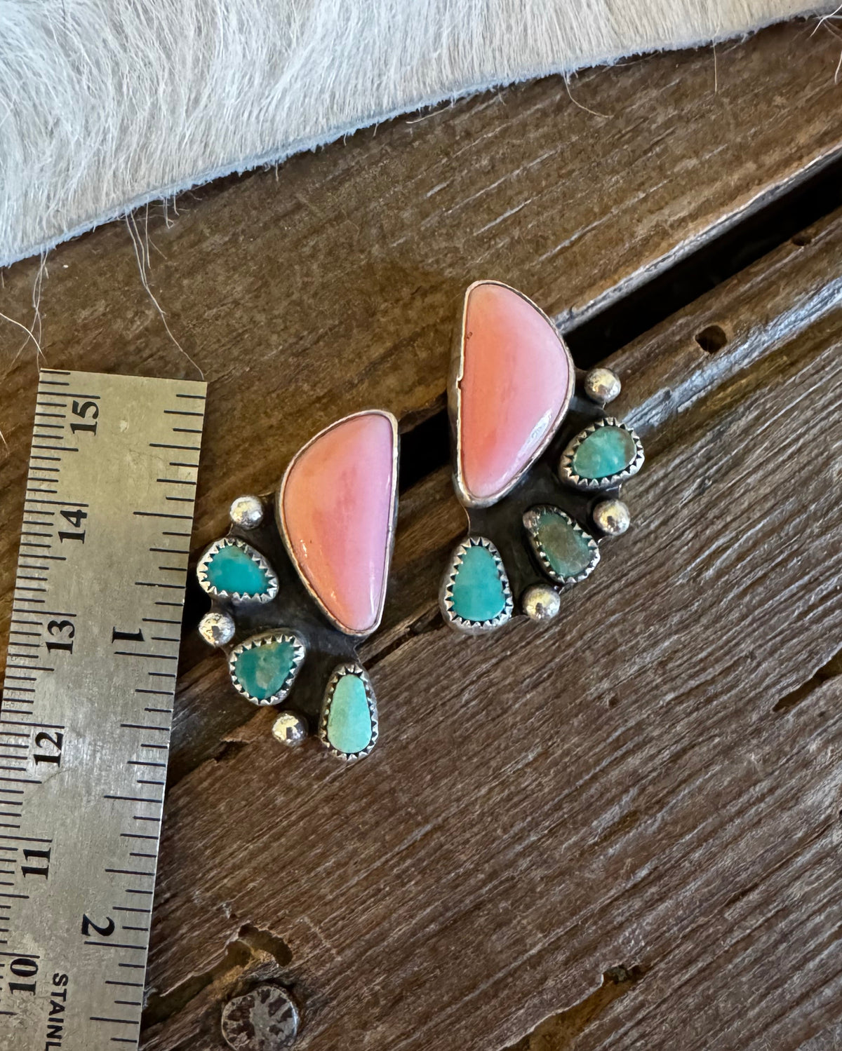 A pair of handmade drop earrings with large pink stone tops, smaller turquoise-blue stones, and silver-toned metal settings, displayed on a rustic wooden surface next to a metal ruler for scale.