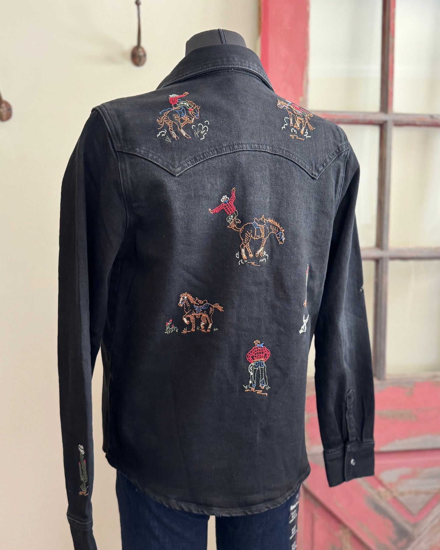 Black denim shirt with embroidered designs on a mannequin against a neutral wall.