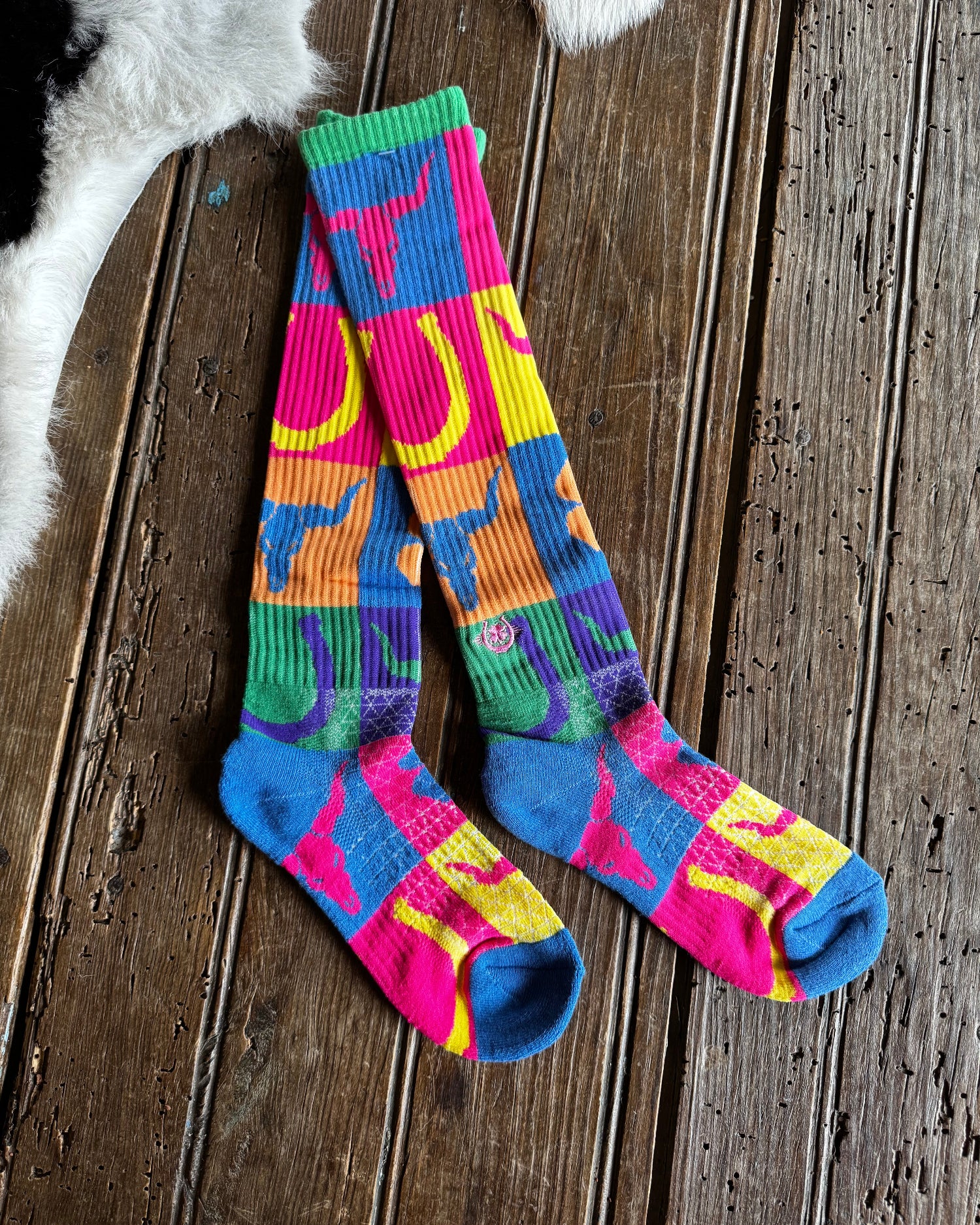 A pair of knee-high socks with a vibrant multicolor patchwork pattern featuring pink, blue, green, yellow, and orange, laid flat on a rustic dark wood plank surface.