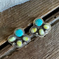 A pair of silver-toned paw-shaped stud earrings featuring turquoise blue round stones and pale yellow-green inlay stones, displayed on a rustic wooden surface with a white faux-fur texture in the background.