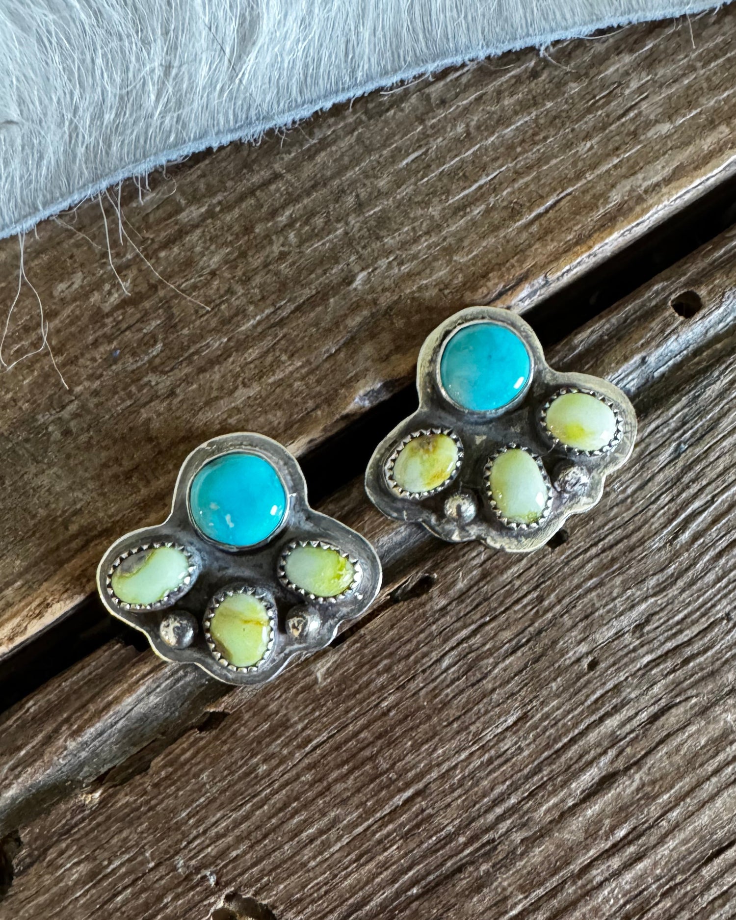 A pair of silver-toned paw-shaped stud earrings featuring turquoise blue round stones and pale yellow-green inlay stones, displayed on a rustic wooden surface with a white faux-fur texture in the background.