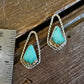 A pair of gold geometric drop earrings with turquoise-colored stone centers, displayed on a rustic wooden surface next to a metal ruler for scale.