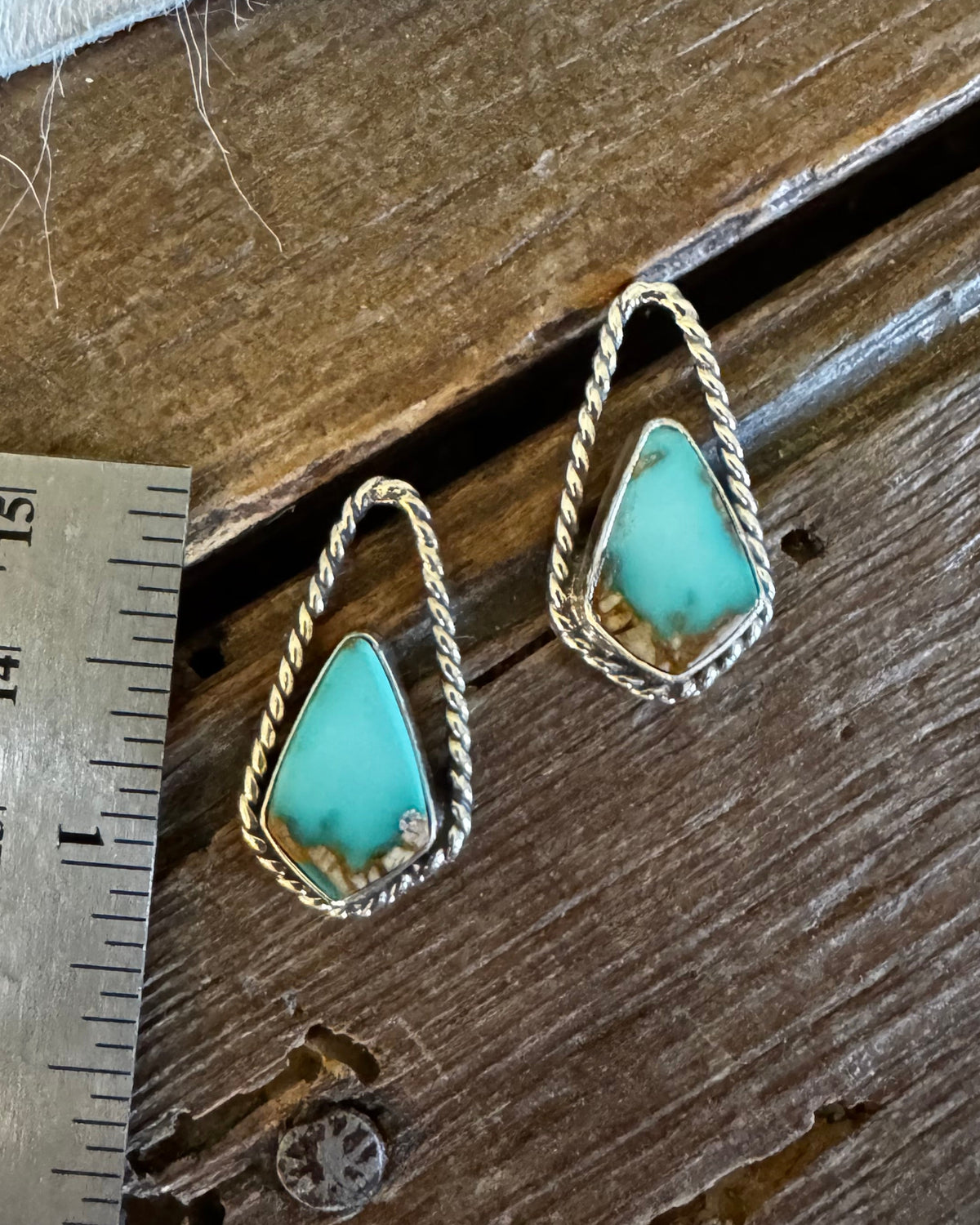 A pair of gold geometric drop earrings with turquoise-colored stone centers, displayed on a rustic wooden surface next to a metal ruler for scale.
