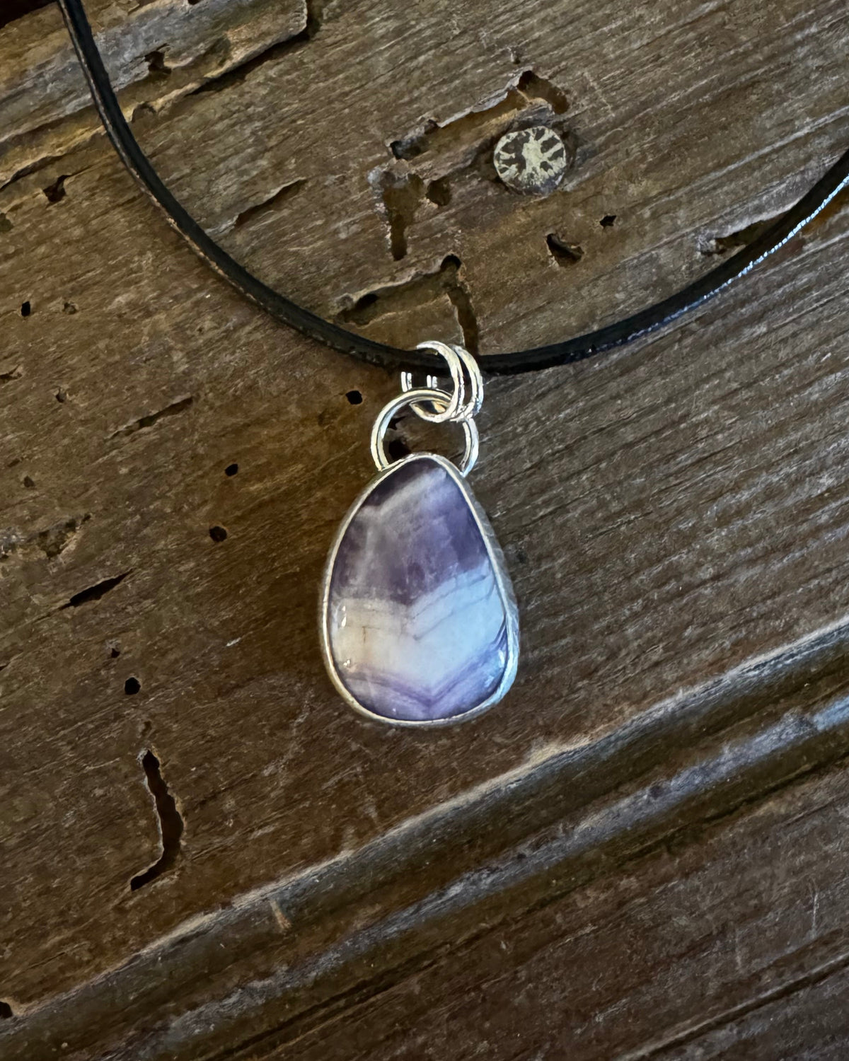 A teardrop-shaped amethyst pendant necklace with a silver bezel and black cord, displayed on a rustic wooden surface.