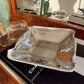 A polished silver rectangular serving tray with a twisted rope border sits on a black book, with two smaller matching glass or clear tray pieces behind it and a decorative mirror in the background.