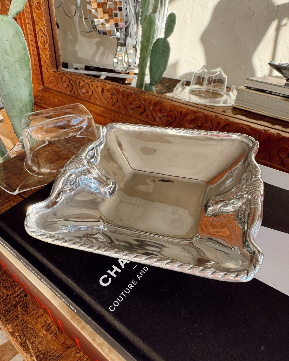 A polished silver rectangular serving tray with a twisted rope border sits on a black book, with two smaller matching glass or clear tray pieces behind it and a decorative mirror in the background.