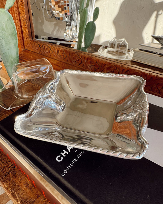 A polished silver rectangular serving tray with a twisted rope border sits on a black book, with two smaller matching glass or clear tray pieces behind it and a decorative mirror in the background.